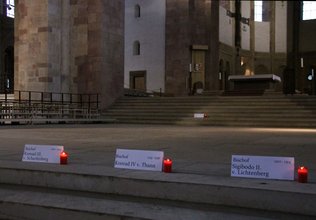 Rote Kerzen stehen auf dem Boden im Königschor des Speyerer Domes vor Namensschildern von im Dom bestatteten Bischöfen